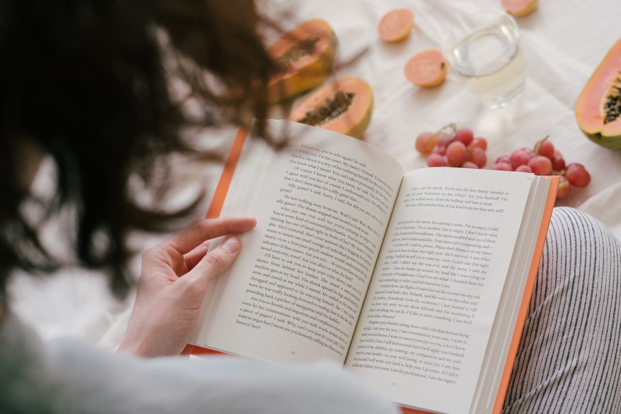 A person enjoys reading a book surrounded by fresh papaya, grapes, and a drink, creating a cozy indoor scene.